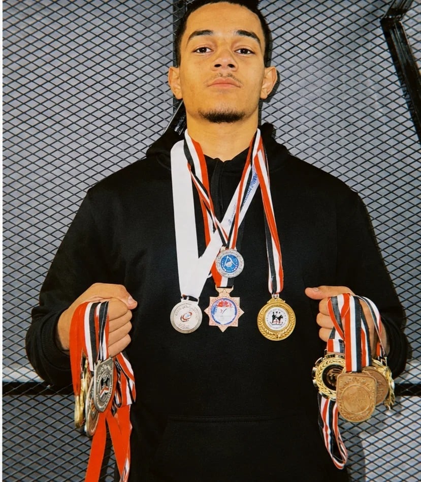 Young man in black jacket displaying multiple medals with orange, white and red ribbons around his neck and held in both hands